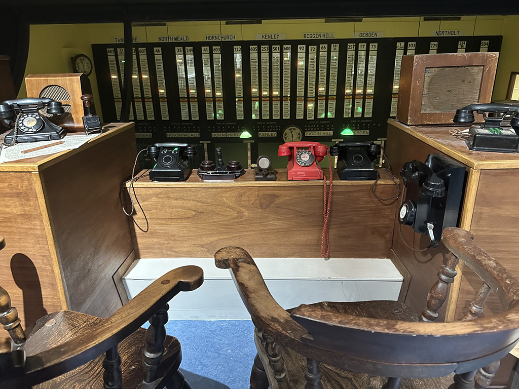 The communications desk inside the Uxbridge Battle of Britain Bunker, featuring vintage rotary telephones, wooden chairs, and a large illuminated status board tracking sector airfields like Biggin Hill and Kenley.